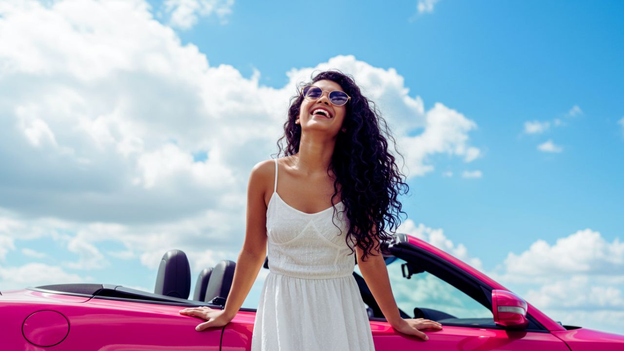 A joyful woman with long, curly dark hair and sunglasses, wearing a white sundress, leaning on a bright pink convertible against a bright blue sky with fluffy white clouds.