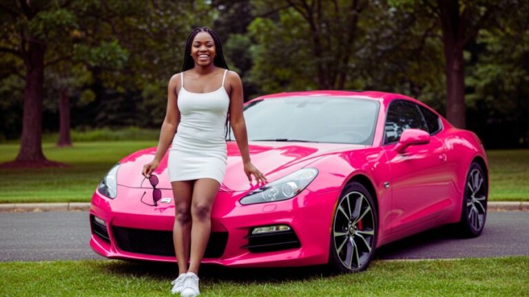A smiling young Black woman in a white dress and white sneakers standing beside a vibrant pink sports car with sunglasses in hand, in an outdoor setting with trees and grass
